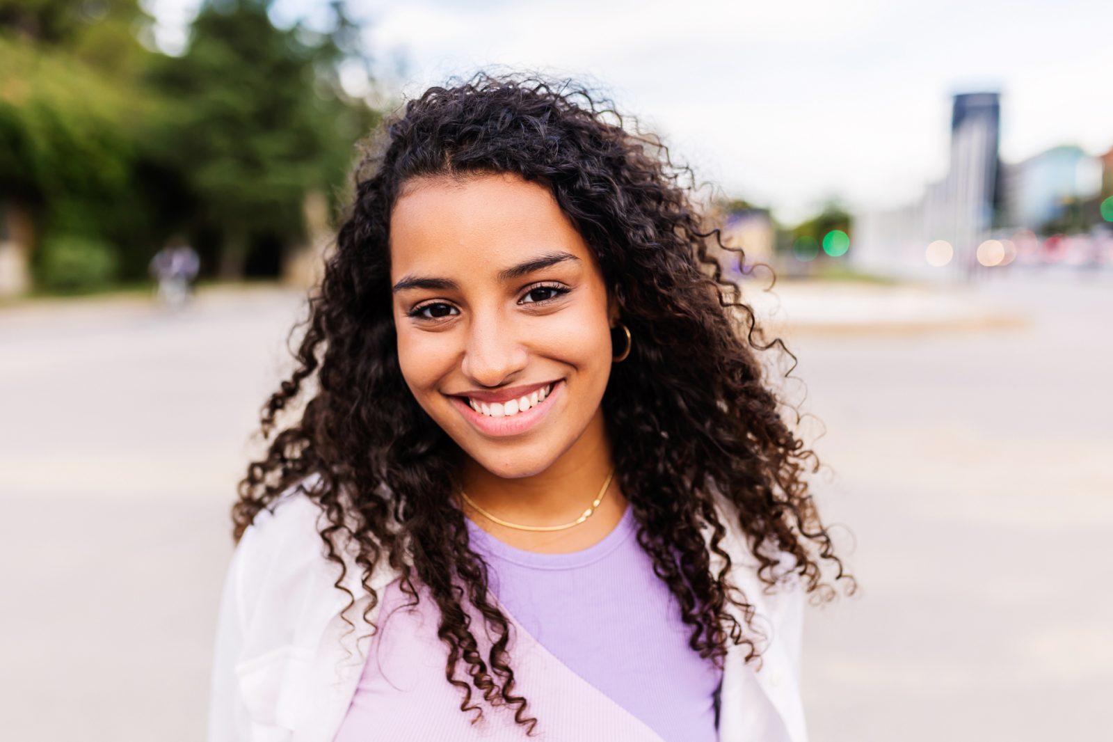 AdobeStock_680745309 Lächelnde Frau mit lockigem, braunen Haar trägt ein lila Oberteil und steht im Freien, Stadt im Hintergrund.