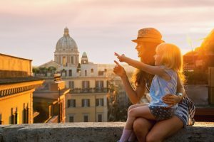 Eine Frau mit Sonnenhut sitzt auf einer Mauer und zeigt mit einem Mädchen auf die Stadt im Sonnenuntergang.