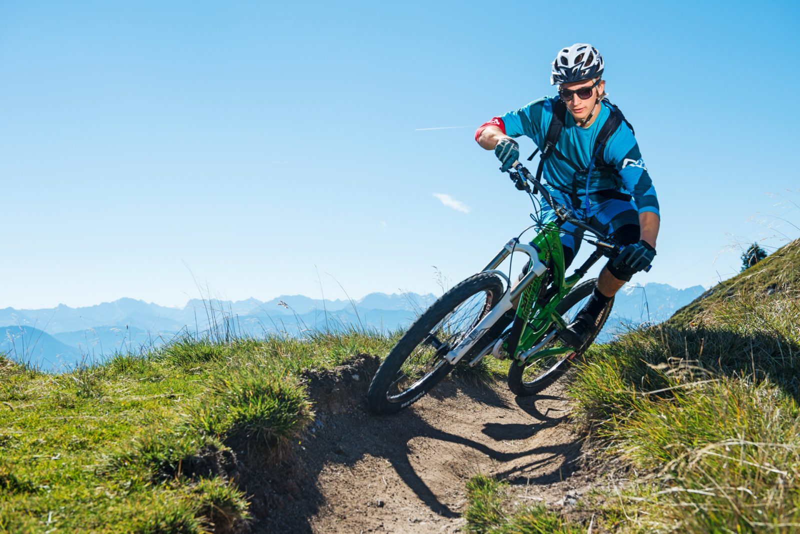 BayernTourismus_JugendTourismus-0485 Mountainbiker auf einem schmalen Pfad, umgeben von grünen Wiesen und Gebirgen unter klarem Himmel.