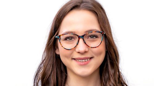 Frau mit langen, braunen Haaren und bunter Brille trägt einen schwarzen Blazer und lächelt vor einem weißen Hintergrund.