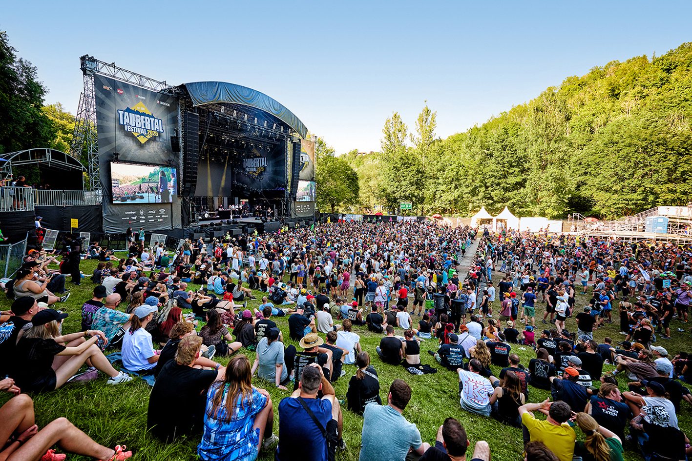 HilltopHoods_(c)FlorianTrykowski_2591957 Festivalbesucher sitzen auf Wiese vor einer Bühne mit großem Monitor, umgeben von Bäumen und einem blauen Himmel.