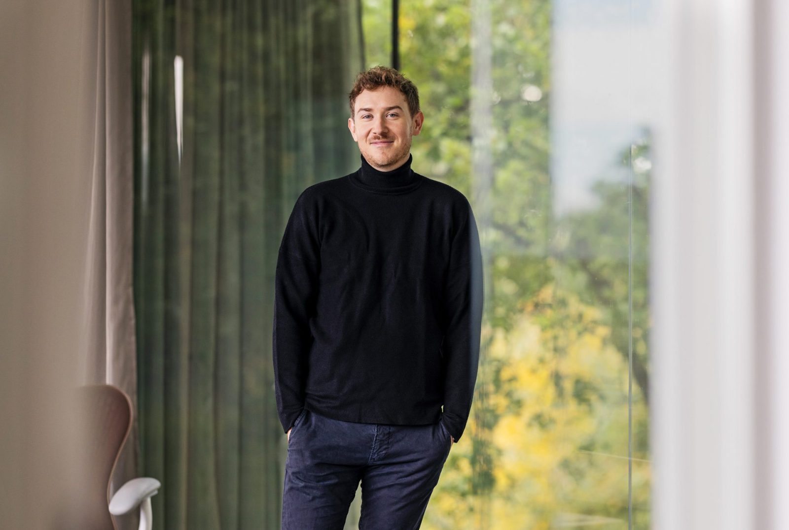 Mann steht in einem modernen Raum mit großen Fenstern und Blick auf grüne Bäume, trägt schwarzen Pullover und Hose.
