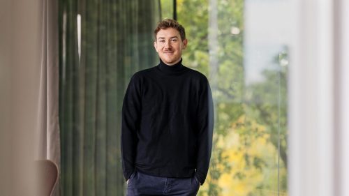 Mann steht in einem modernen Raum mit großen Fenstern und Blick auf grüne Bäume, trägt schwarzen Pullover und Hose.