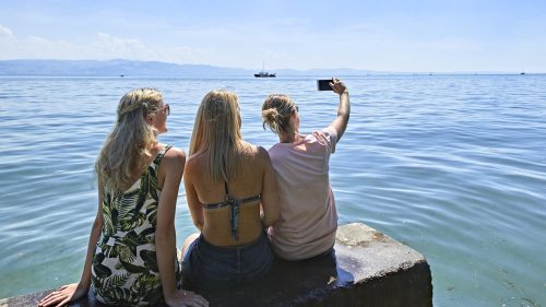 Drei Frauen sitzen am Ufer eines Sees und machen ein Selfie, während ein Boot in der Ferne fährt.
