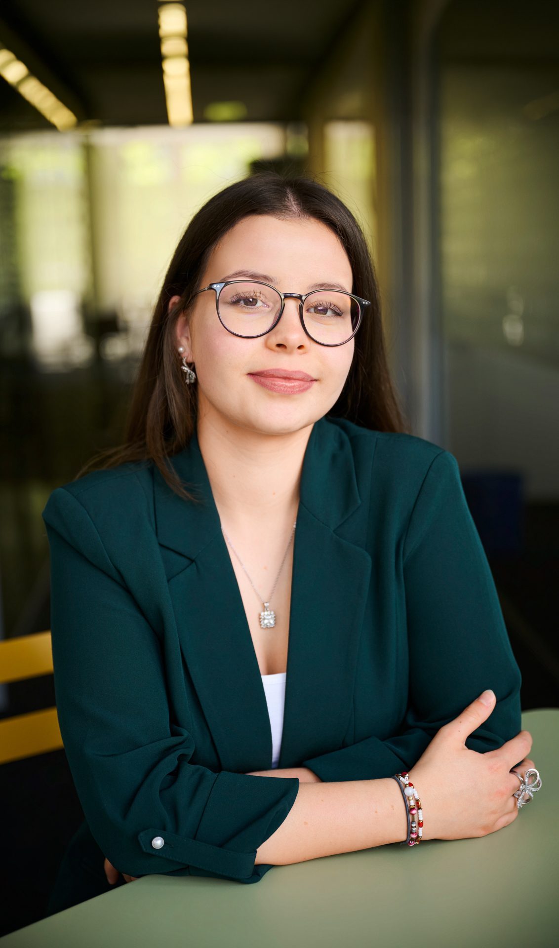 bay-tm_corinna_schachtner_0055 Porträt einer jungen Frau mit Brille, grünem Blazer und Schmuck, lächelt vertrauensvoll an einem Tisch.
