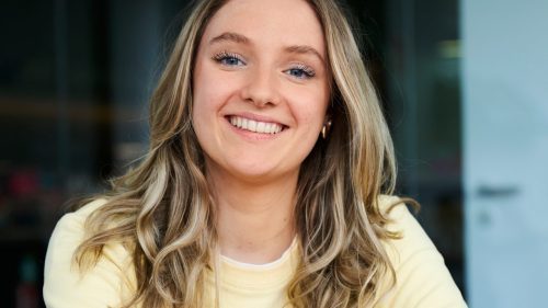 Eine lächelnde Frau mit langem, blondem Haar sitzt an einem roten Tisch und trägt einen gelben Pullover.