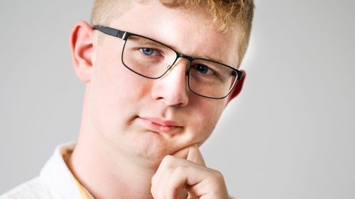 Junger Mann mit Brille und gestreiftem Hemd, der nachdenklich mit der Hand an seinem Kinn steht.