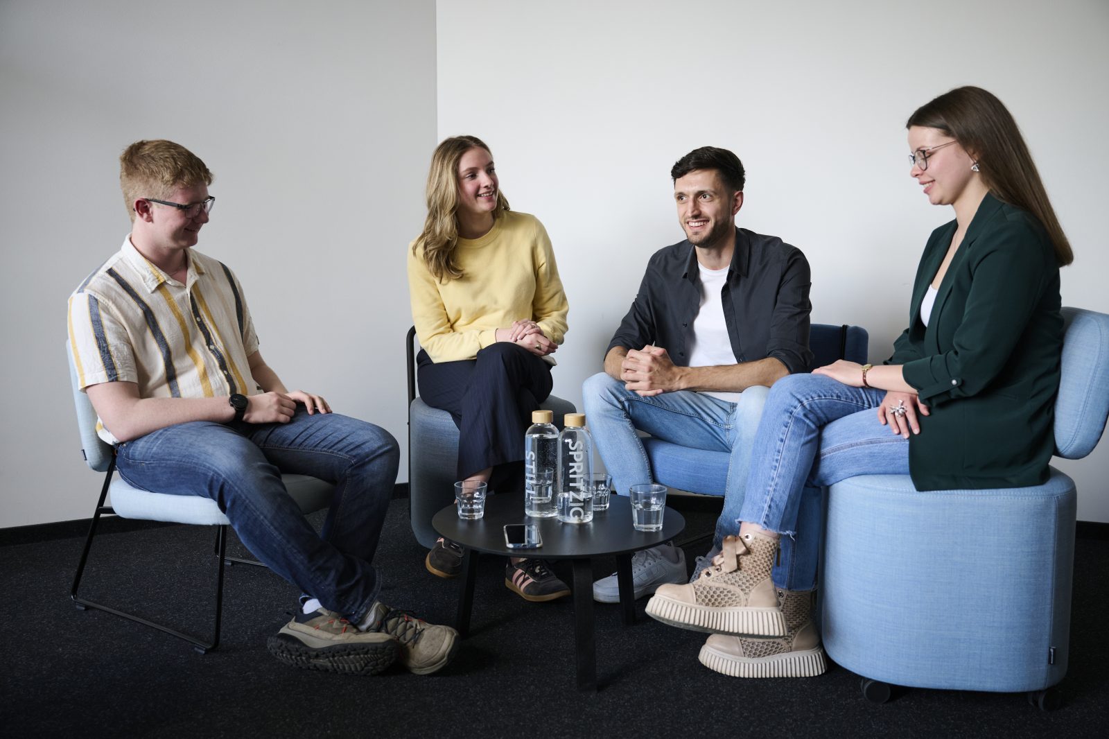 Vier Personen sitzen in moderner Büroeinrichtung bei einem Interview, Wasserflaschen und Gläser auf Tisch.