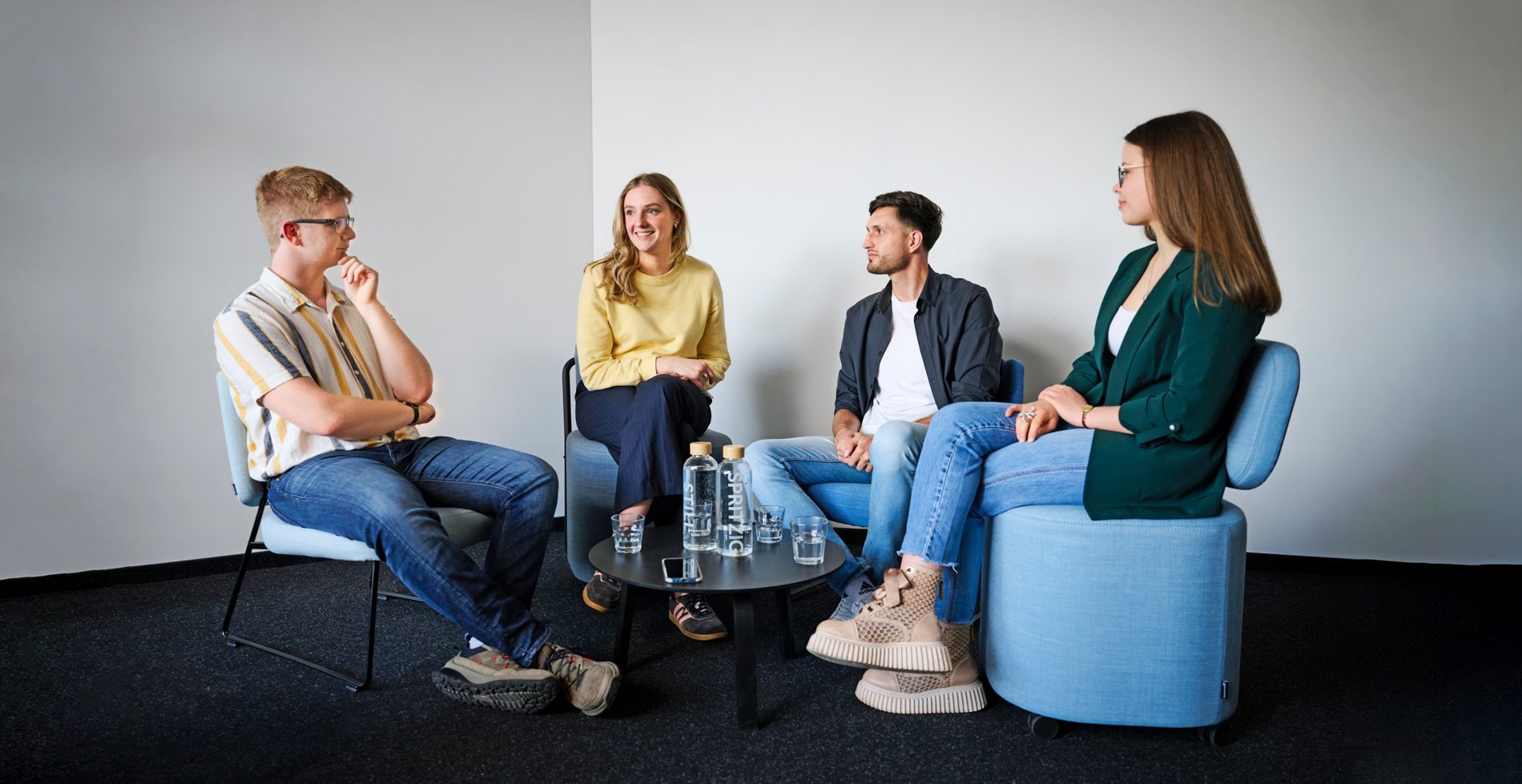 bay-tm_interview_0309_a Vier Personen sitzen in einem hellen Raum zusammen. Sie diskutieren an einem Tisch mit Wasserflaschen und Gläsern.