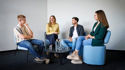Vier Personen sitzen in einem hellen Raum zusammen. Sie diskutieren an einem Tisch mit Wasserflaschen und Gläsern.