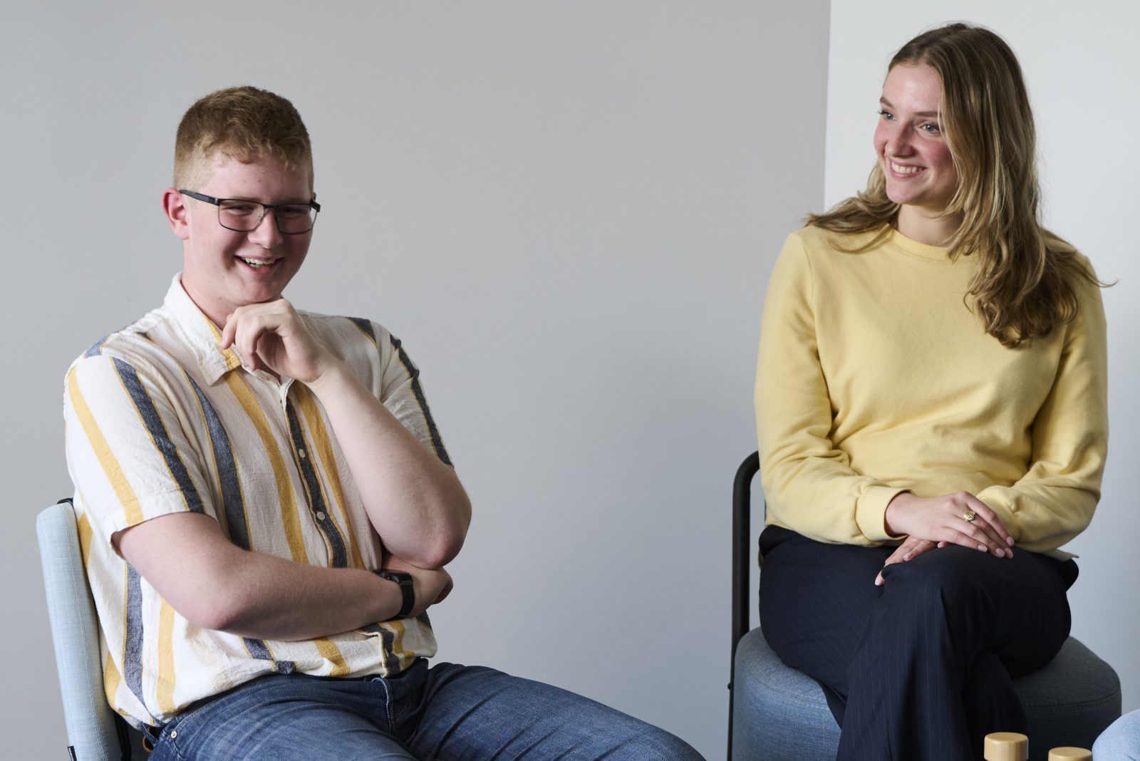 bay-tm_interview_0383_quer Zwei junge Menschen sitzen in einem Gespräch. Ein Junge mit Brille und gestreiftem Hemd lächelt, ein Mädchen in gelbem Pullover sitzt daneben.