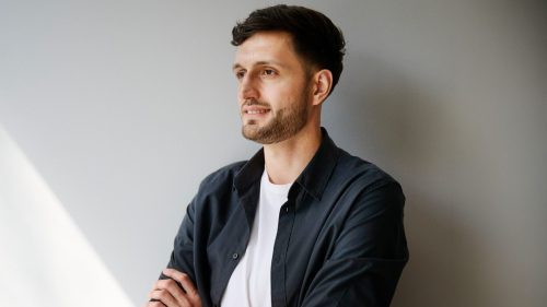 Mann mit kurzem Haar in schwarzer Jacke und weißem T-Shirt, verschränkt die Arme vor einer grauen Wand.