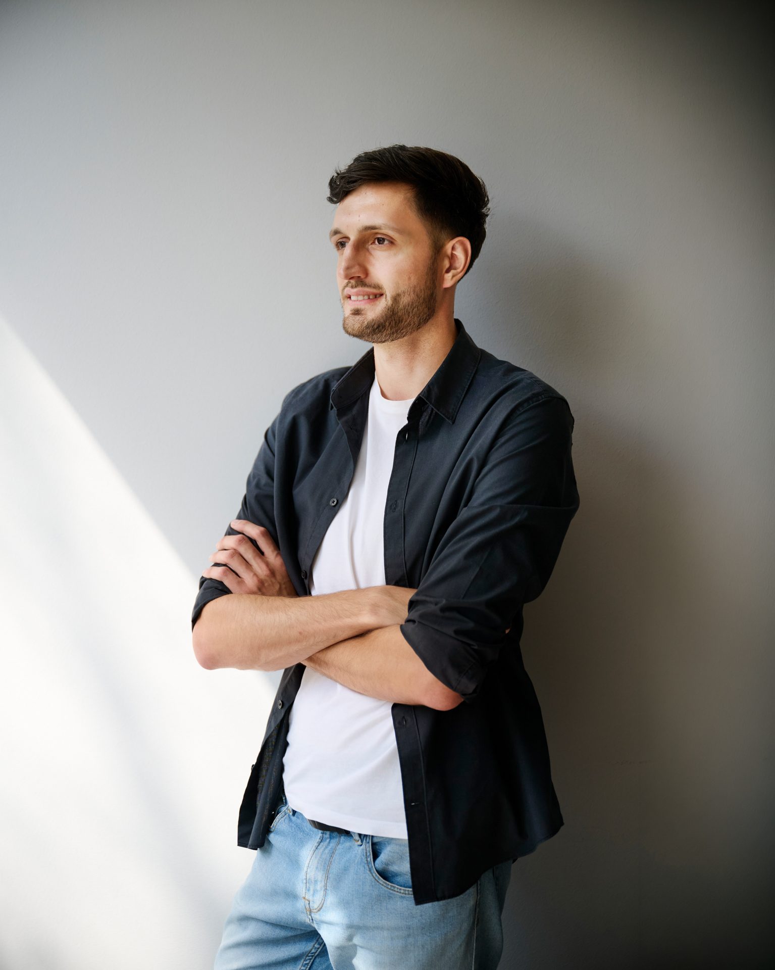 bay-tm_martin_faethe_0014 Mann mit kurzem Haar in schwarzer Jacke und weißem T-Shirt, verschränkt die Arme vor einer grauen Wand.