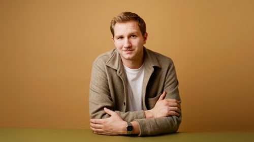 Mann mit kurzem, blonden Haar sitzt an einem Tisch, trägt ein weißes T-Shirt mit beiger Jacke und blickt in die Kamera.